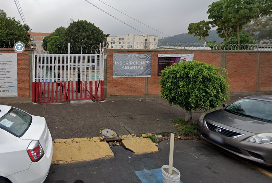 Fotografía en la que se muestra la entrada del plantel, conformada por una reja de color blanco con rojo y una rampa de acceso. Del lado izquierdo y derecho de la reja hay muros hechos de ladrillo. Sobre uno de los muros hay una lona que dice CECATI 12, INSCRIPCIONES ABIERTAS.